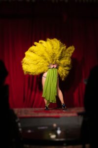 A DC burlesque fan dancer Isabelle Epoque faces away from the camera. Her Chartreuse feather fans are spread behind her back showing off her legs